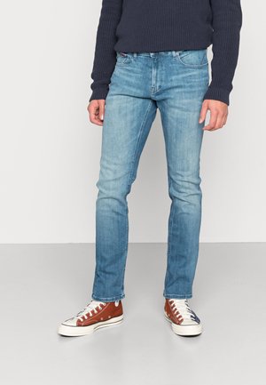 Personne portant un jean slim bleu clair, un pull bleu marine et des baskets montantes bicolores marron et bleu marine, debout sur un sol gris.