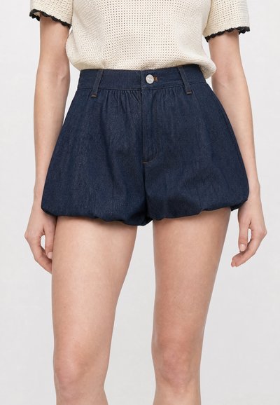 Femme portant un short ballon en denim foncé taille haute avec fermeture bouton et zip, associé à un top en maille blanc à manches courtes.