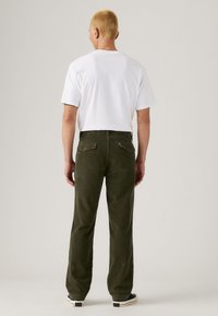 Olivgrüne Cordhosen mit Gesäßtaschen, kombiniert mit einem schlichten weißen T-Shirt. Modell mit dem Rücken zur Kamera. Gummisohlen-Schuhe sind sichtbar.