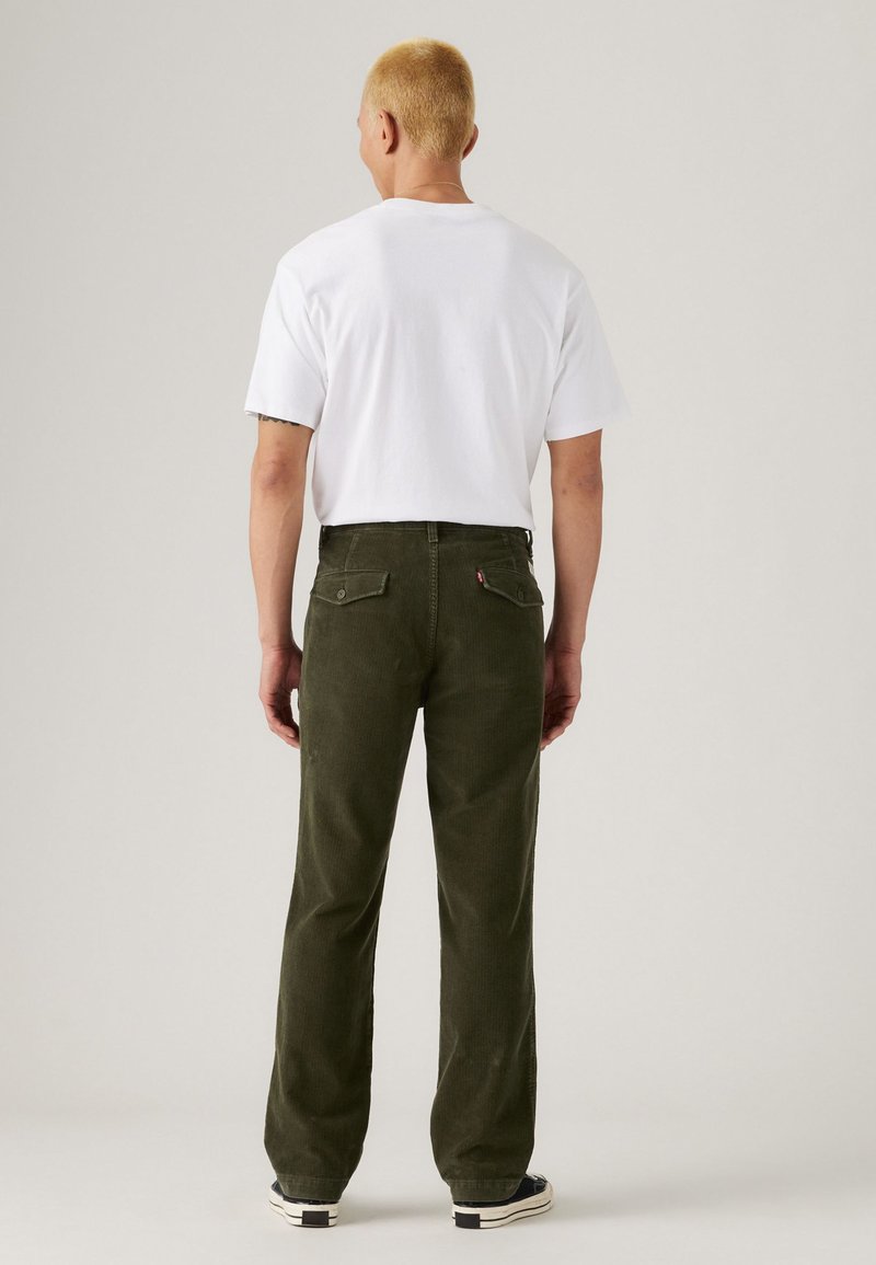 Olive green corduroy pants with back pockets, paired with a plain white t-shirt. Model facing away from the camera. Rubber-soled shoes visible.