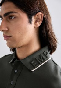 Σκούρο πράσινο πόλο μπλουζάκι με υφή στον γιακά που έχει αναγλυφές το "EMP". Διαθέτει λευκή αντίθεση στην περιφέρεια και μαύρα κουμπιά στη στεφάνη.