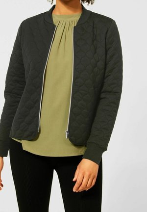 Femme portant une veste matelassée zippée vert foncé par-dessus un chemisier vert clair et un pantalon noir, debout devant un fond uni.