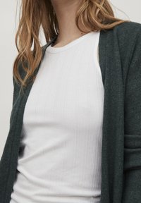 Mörkgrön cardigan i texturerat material, lager på lager över en åtsittande vit ribbad linne. Fokus på tyg och färgkontrast.