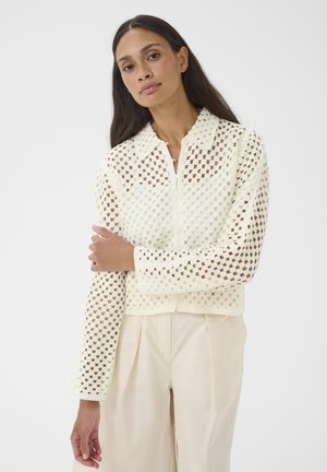 Femme aux longs cheveux foncés portant un haut en crochet crème à manches longues et un pantalon beige taille haute, debout devant un fond uni.
