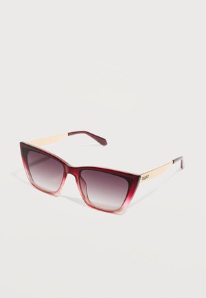 Katí oči sluneční brýle s červenými gradientními obroučkami, zlatými kovovými stranami a tmavými tónovanými čočkami, ležící na světlém pozadí.