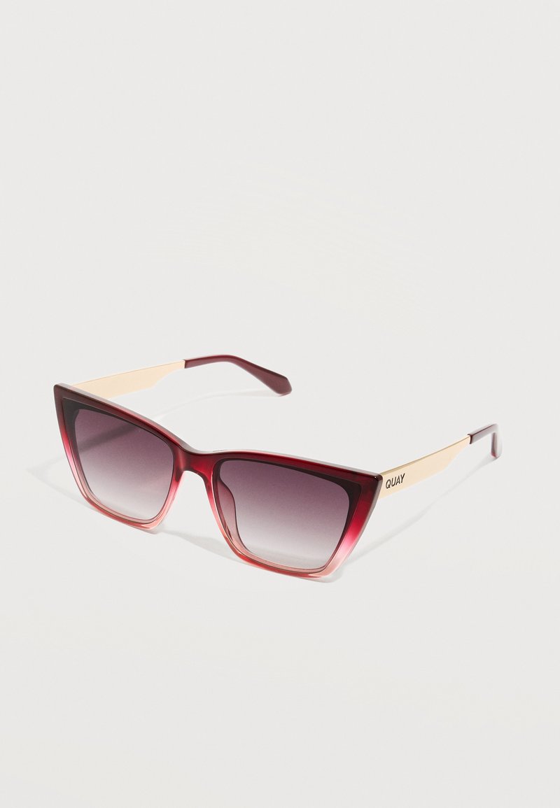 Γυαλιά ηλίου με σκελετό σε σχήμα γάτας σε κόκκινη διαβάθμιση, χρυσά μεταλλικά μπράτσα και σκουρόχρωμους φακούς, τοποθετημένα σε ανοιχτό φόντο.