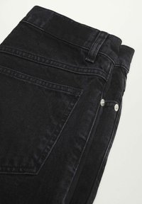 Gros plan sur un jean en denim noir montrant une poche arrière, une passants de ceinture et des rivets métalliques sur un fond clair.
