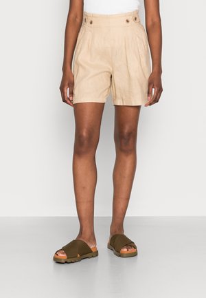 Person iført beige højtaljede plisserede shorts med knapdetaljer og olivengrønne slidesandaler på hvid baggrund.