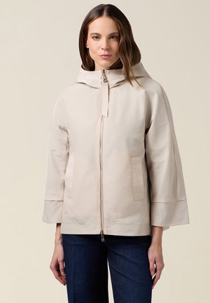 Frau trägt eine beige Reißverschlussjacke mit hohem Kragen und weiten Ärmeln, kombiniert mit dunkelblauen Jeans, vor einem schlichten hellen Hintergrund.