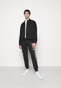 Chaqueta de mezclilla negra con bolsillos frontales sobre una camiseta blanca, combinada con joggers negros que presentan un logo y acentos blancos, completada con zapatillas blancas.