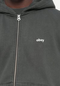 Svetrová mikina na zips v tmavo sivom materiáli, s malým bielym logom "obey" v hornej ľavej časti a jemným, textúrovaným vzhľadom.