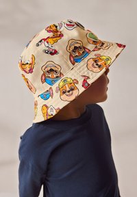 Kind trägt einen beigen Fischerhut mit bunten Cartoon-Hundegesichtern und ein dunkelblaues Langarmshirt, seitlich betrachtet vor einem schlichten Hintergrund.