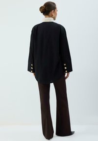 Camicia nera a maniche lunghe con colletto alto bianco, con dettagli di bottoni dorati sulle maniche, abbinata a pantaloni marroni a zampa d'elefante.