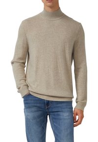 Mann in beigem, geripptem Rollkragenpullover und blauen Jeans, mit einer Hand in der Tasche vor weißem Hintergrund.