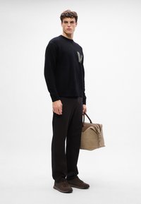 Suéter negro de punto con un logo 'M' texturizado, combinado con pantalones oscuros. Bolso marrón claro con acentos más oscuros, zapatos marrones, superficie lisa.