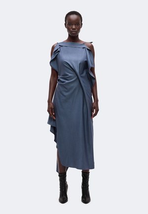 Femme portant une robe asymétrique bleue avec des détails volantés aux épaules dénudées et des bottines noires, debout devant un fond uni.