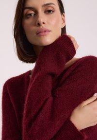 Burgundowa sweter wykonany z miękkiego, puszystego materiału, z długimi rękawami i luźnym krojem, z ribbymi mankietami i dekoltem w kształcie V.