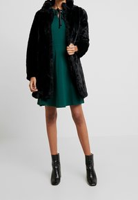 Svart fuskpälsjacka med huva, långa ärmar och en avslappnad passform, lager på lager över en grön klänning. Svarta ankelskor fulländar outfiten.