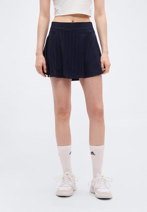 Femme portant un haut court blanc, une jupe de tennis plissée marine, des chaussettes blanches Adidas et des baskets blanches, debout devant un fond uni.