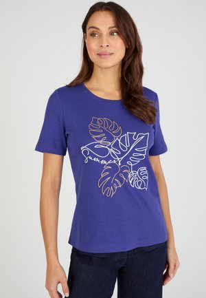 Vrouw draagt een paarse shirt met korte mouwen met witte en gouden tropische bladcontouren en het woord "summer" op de voorkant, gecombineerd met een donkere spijkerbroek.