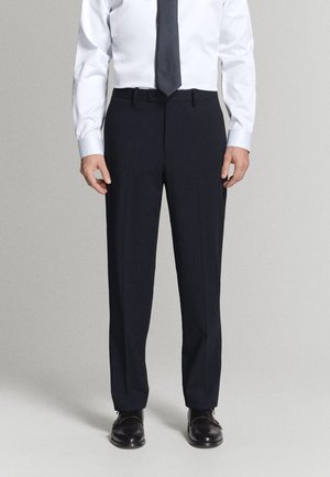 Mężczyzna ubrany w czarne eleganckie spodnie, czarne wizytowe buty, białą koszulę z długim rękawem i ciemny krawat, stoi na jednolitym tle.