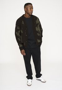 Dunkelgrüner und schwarzer gemusterter Cardigan mit durchgehender Knopfleiste, getragen über einem schwarzen Shirt. Kombiniert mit schwarzen Hosen und weißen Schuhen.