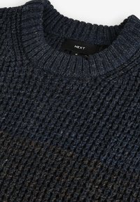 Pulover tricotat navy cu guler rotund ribat. Prezintă o suprafață texturată și variații subtile de culoare, cu un eticheta de marcă neagră vizibilă.