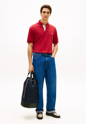 Młody mężczyzna w czerwonej koszulce polo, niebieskich dżinsach i czarnych mokasynach, trzymający dużą granatową plecioną torbę na ramię z czarnymi uchwytami.