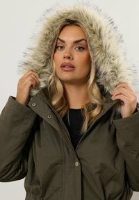 Olivgrön puffjacka med foder av syntetisk päls i huooden, med dragkedja och snapknappar. Slät textur, rymlig passform.