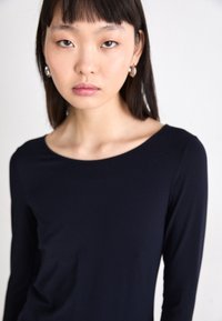 Marineblauwe longsleeve top met een brede bootnek, gemaakt van zacht materiaal. Model draagt zilveren oorbellen in de vorm van een ring. Effen achtergrond.