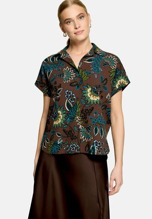 Femme portant une chemise marron à manches courtes à motifs floraux avec des touches vertes, bleues et jaunes, associée à une jupe marron foncé.