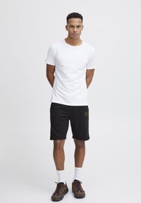 Camiseta blanca de algodón combinada con pantalones cortos negros con un logo. El modelo lleva calcetines blancos y zapatillas de deporte marrones. Diseño simple y casual.