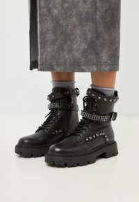 Botas de combate de cuero negro con acentos de tachuelas, acabado texturizado y suela negra gruesa. Presentan correas y ojales decorativos de rhinestone.