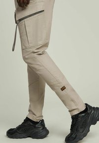 Pantaloni cargo beige con molte tasche, dettagli con zip e etichetta a patch; abbinati a sneakers nere chunky.