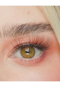 Nahaufnahme eines Auges mit haselnussbraunem Iris, langen schwarzen Wimpern, subtiler rosa Lidschatten und deutlichem Licht, das im Pupil reflektiert wird.
