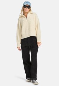 Cremfarbener Fleece-Pullover mit einem halben Reißverschluss, kombiniert mit schwarzen Cordhosen und grauen Leinenschuhen. Ergänzt mit einer hellblauen Kappe.