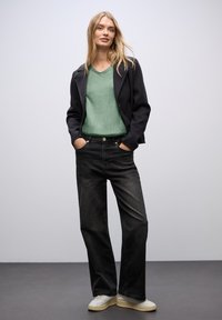 Kobieta z długimi blond włosami, ubrana w czarną kurtkę, zielony top w serek, czarne szerokie dżinsy, białe tenisówki, stoi z rękami w kieszeniach.