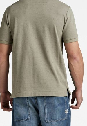 Homme portant une chemise beige à manches courtes et un short en jean bleu vu de dos, avec un tatouage visible sur l'avant-bras droit.