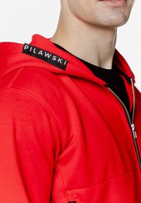 Czerwona bluza z kapturem na zamek błyskawiczny o gładkiej fakturze, z czarną metką marki "PILAWSKI" na lewym ramieniu. Materiał polyesterowy.