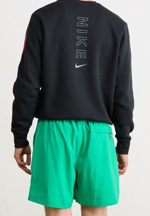 Persoon die een zwart shirt met lange mouwen draagt met verticaal lichtblauw "NIKE"-logo en groene shorts, van achteren gezien tegen een effen achtergrond.