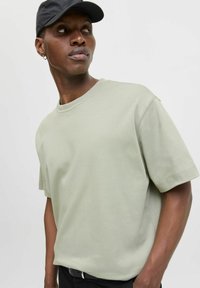 Tricou din bumbac verde deschis, cu mâneci scurte, decolteu rotund și o croială lejeră. Este purtat cu o șapcă neagră și pantaloni de culori închise.