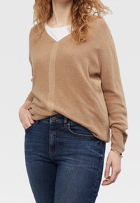 Mujer con un suéter de cuello en V color beige sobre una blusa blanca y jeans azul oscuro, de pie con una mano en el bolsillo frente a un fondo liso.