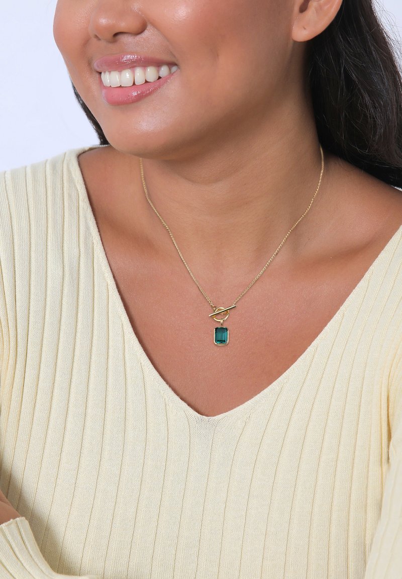 Collier en or arborant une pierre précieuse verte rectangulaire, suspendu à un design de maillons torsadés, porté sur un pull à côtes léger.