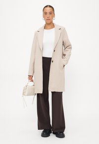 Beige wollen jas met notch-lapels, twee knopen aan de voorkant en zijzakken; gecombineerd met bruine wijde broek en zwarte sneakers.