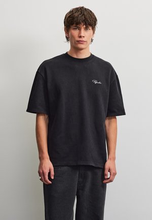 Giovane uomo con capelli castani corti che indossa una maglietta nera a coste larga e pantaloni scuri su uno sfondo chiaro uniforme.