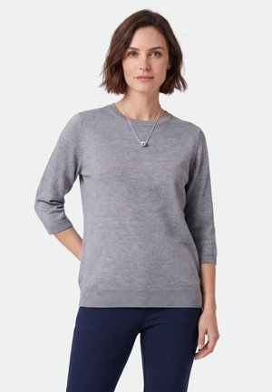 Kvinde iført en lysegrå sweater med trekvart-ærmer og mørke bukser, stående med en hånd bag ryggen og et sølvhalskæde.