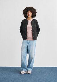 Giacca nera trapuntata con bottoni a pressione, indossata sopra una felpa rosa chiaro con scritta bianca, abbinata a jeans blu larghi e scarpe da ginnastica bianche.