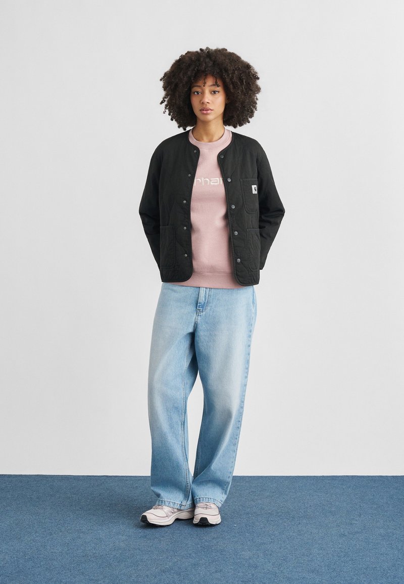 Zwarte doorgestikte jas met drukknoopsluiting, gedragen over een lichtroze sweatshirt met witte tekst, gecombineerd met losse blauwe jeans en witte sneakers.