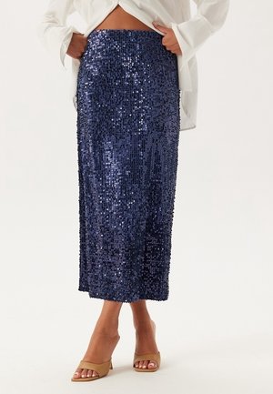 Jupe midi bleu marine à sequins portée avec des talons ouverts beige et un chemisier blanc maintenu ouvert à la taille.
