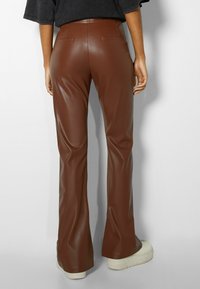 Pantaloni a zampa d'elefante in finta pelle marrone con una texture liscia, due tasche posteriori e una silhouette aderente, abbinati a sneakers bianche con zeppa.